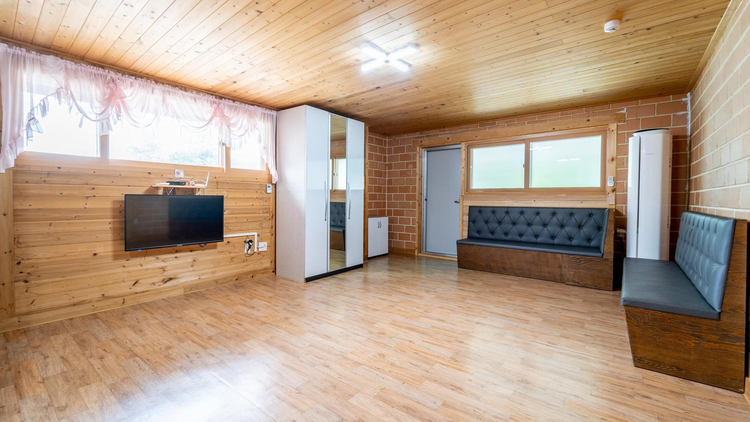 A room with wooden walls and ceiling, a television mounted on the wall, a bench, and a window with curtains.