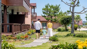 A family walking on a pathway beside a building with a wooden deck and a garden with various plants and flowers. - Jeju Sigan Yeohaeng Huyang Pension (Jeju City)