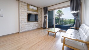 A spacious living room with a wooden floor, a sofa, a coffee table, a flat-screen TV mounted on a brick wall, and a balcony with a view of palm trees. - Seogwipo Narsha Resort (Seogwipo)