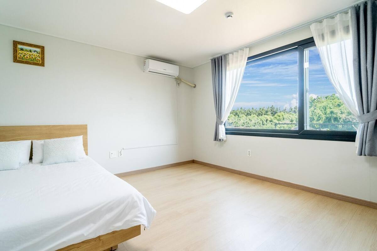 Un dormitorio con cama, aire acondicionado, ventana con cortinas y vista a zonas verdes.