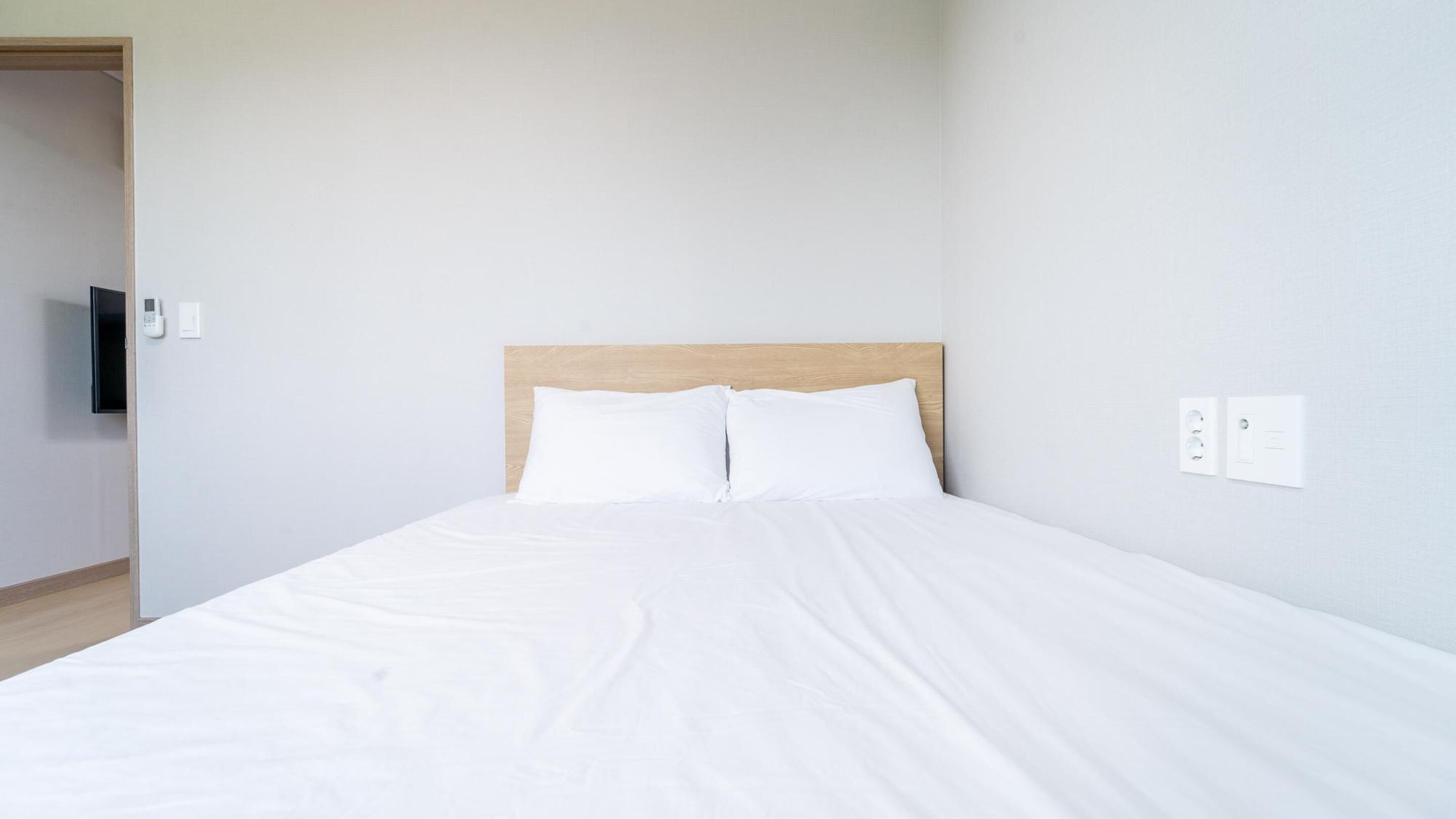 A neatly made bed with white linens in a room with light-colored walls and a wooden headboard.