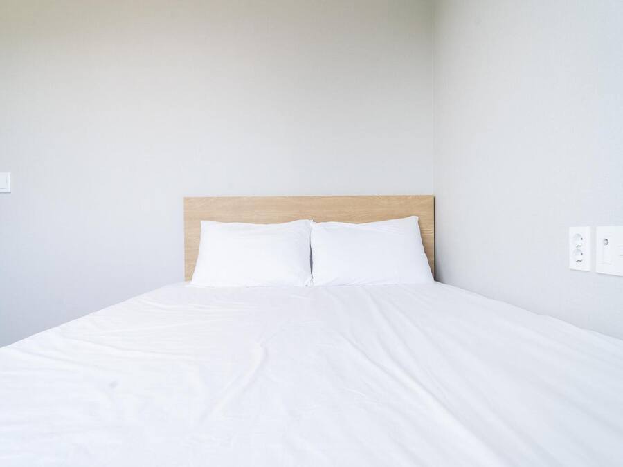 A neatly made bed with white linens in a room with light-colored walls and a wooden headboard.
