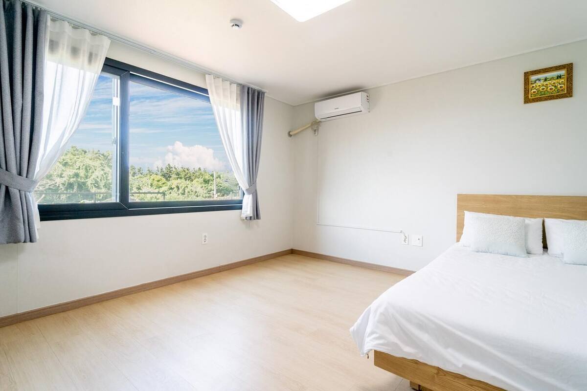 Un dormitorio con cama, ventana con cortinas, equipo de aire acondicionado y un cuadro colgado en la pared.