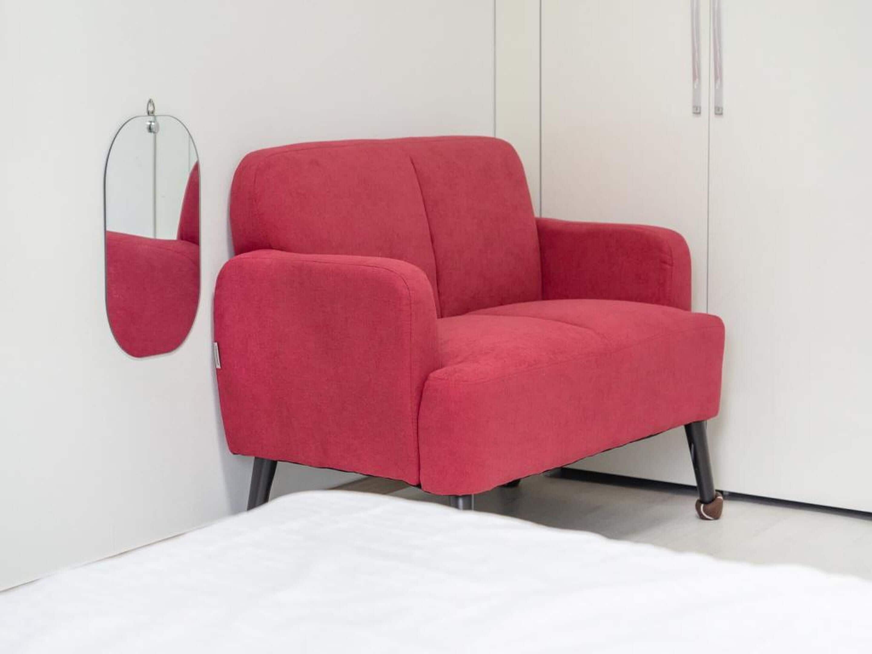 A red armchair with wheels in a minimalist room with white walls and a white bed.