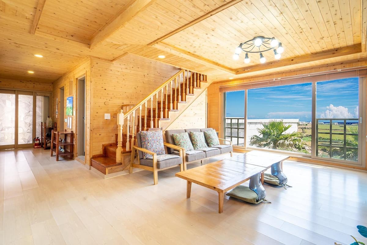 A spacious living area with a wooden staircase, a sofa set, and a large window offering a view of the outdoors.