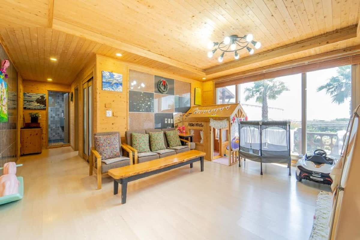 A cozy room with a wooden ceiling, a sofa with cushions, a bench, a playpen, and a view of palm trees outside.