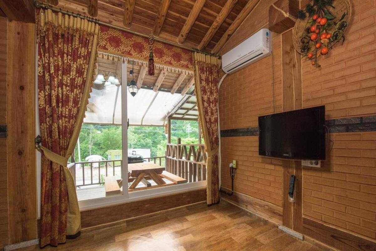 A wooden interior with a TV mounted on the wall, a wooden bench outside, and a view of greenery through large windows.