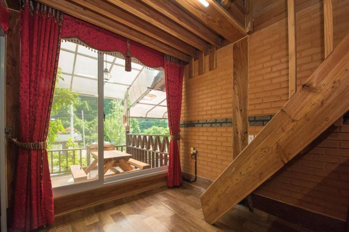 A wooden interior with a wooden staircase, a wooden table, and a window with red curtains.