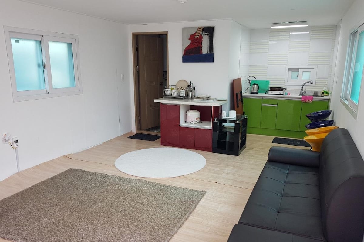 Un salon avec un canapé, un tapis rond, un coin cuisine doté d’un plan de travail vert, et une porte donnant sur une autre pièce.