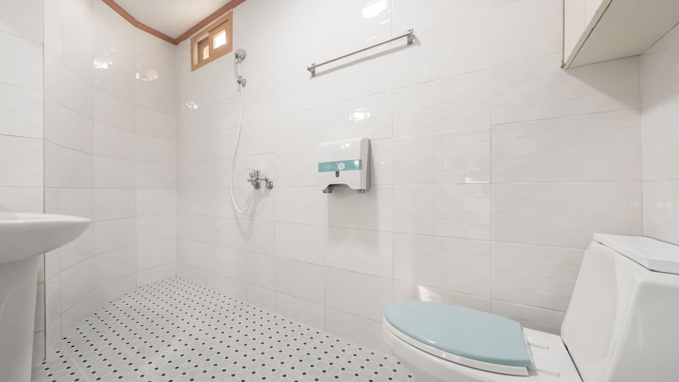 Une salle de bain carrelée de carreaux blancs, des toilettes avec un abattant bleu clair, une douche et un sèche-mains.
