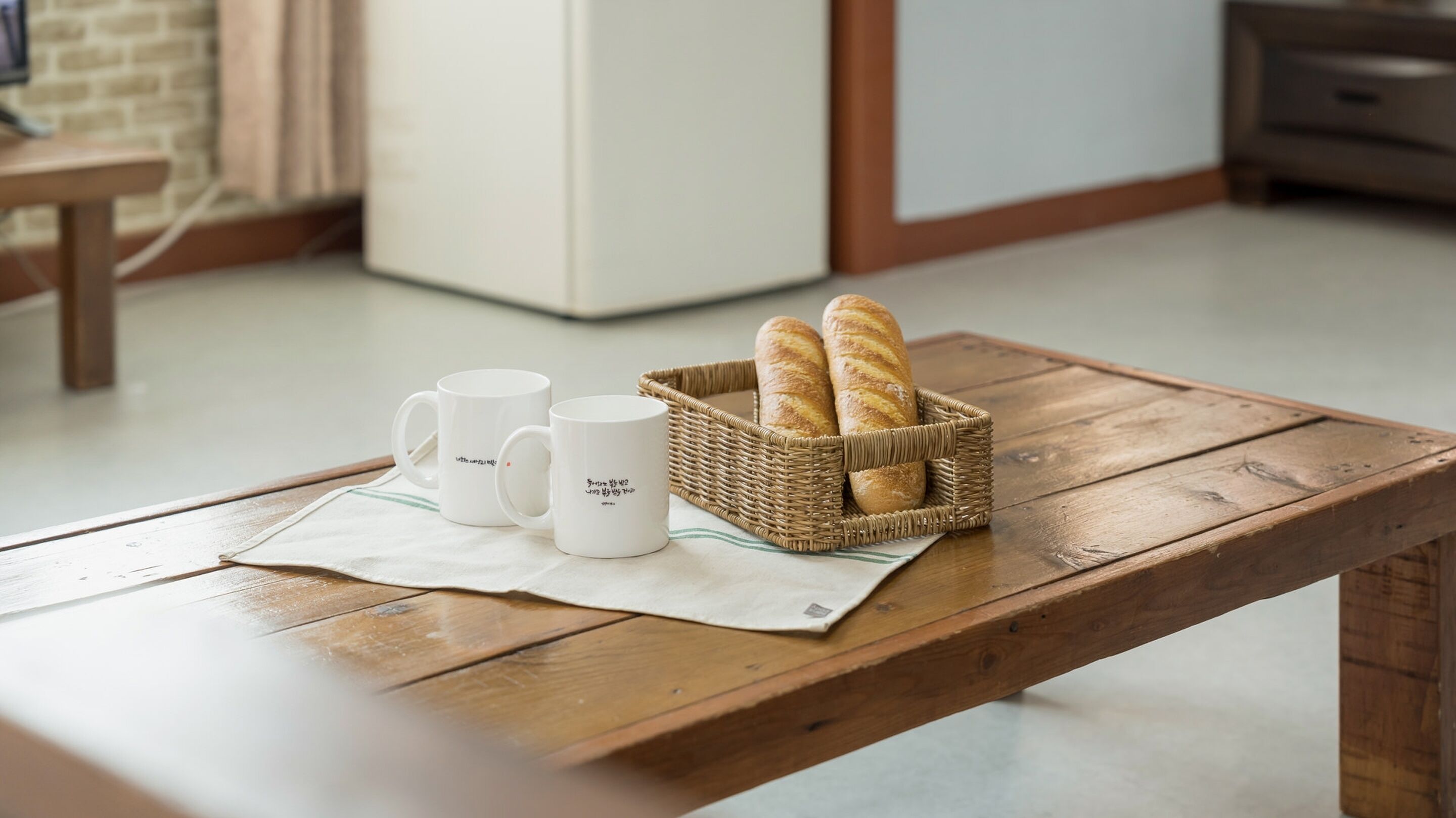A wooden table with two white mugs and a wicker basket containing two baguettes.