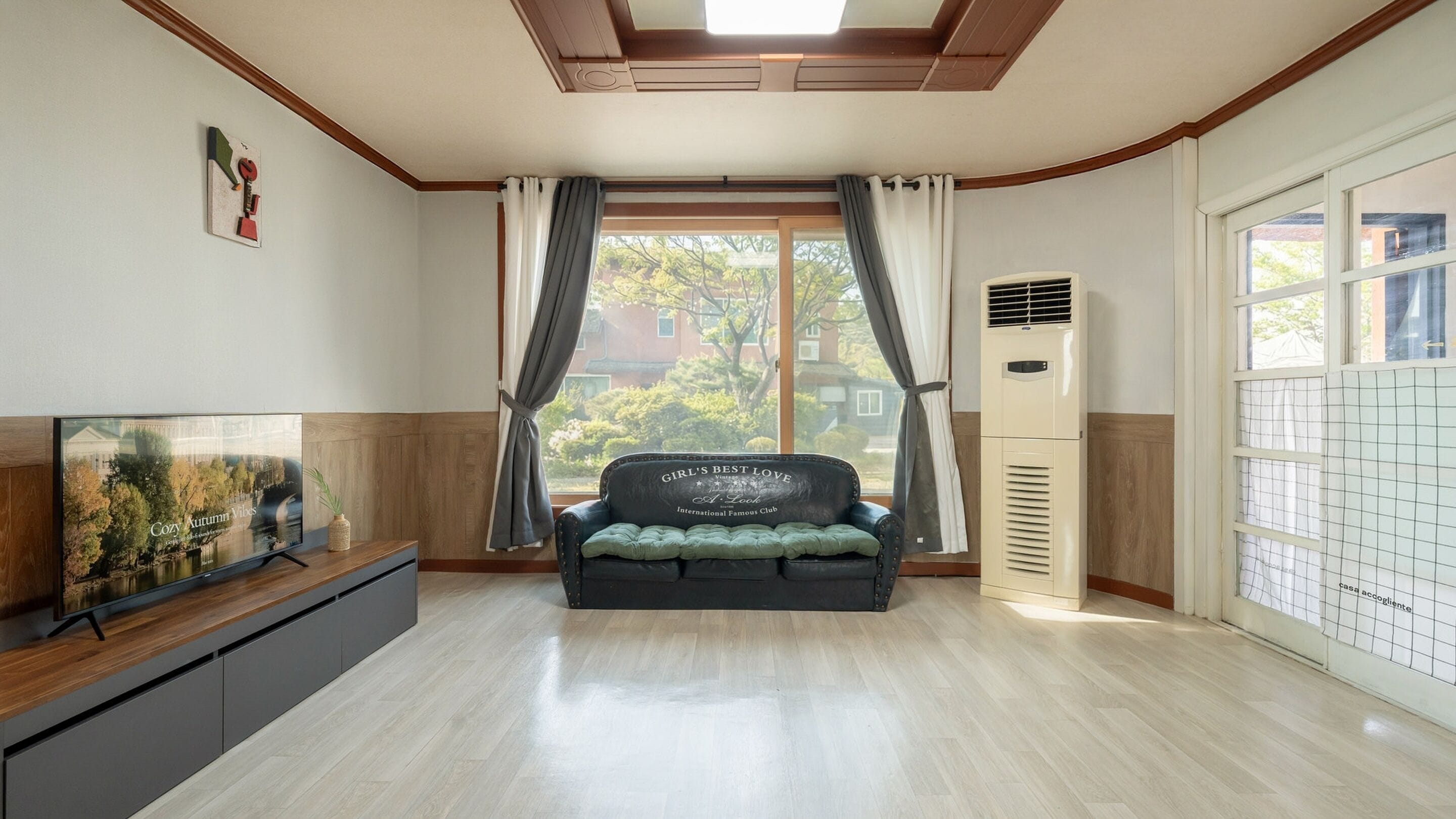 A room with a sofa bed, a TV, a window with curtains, and a wall-mounted air conditioner.