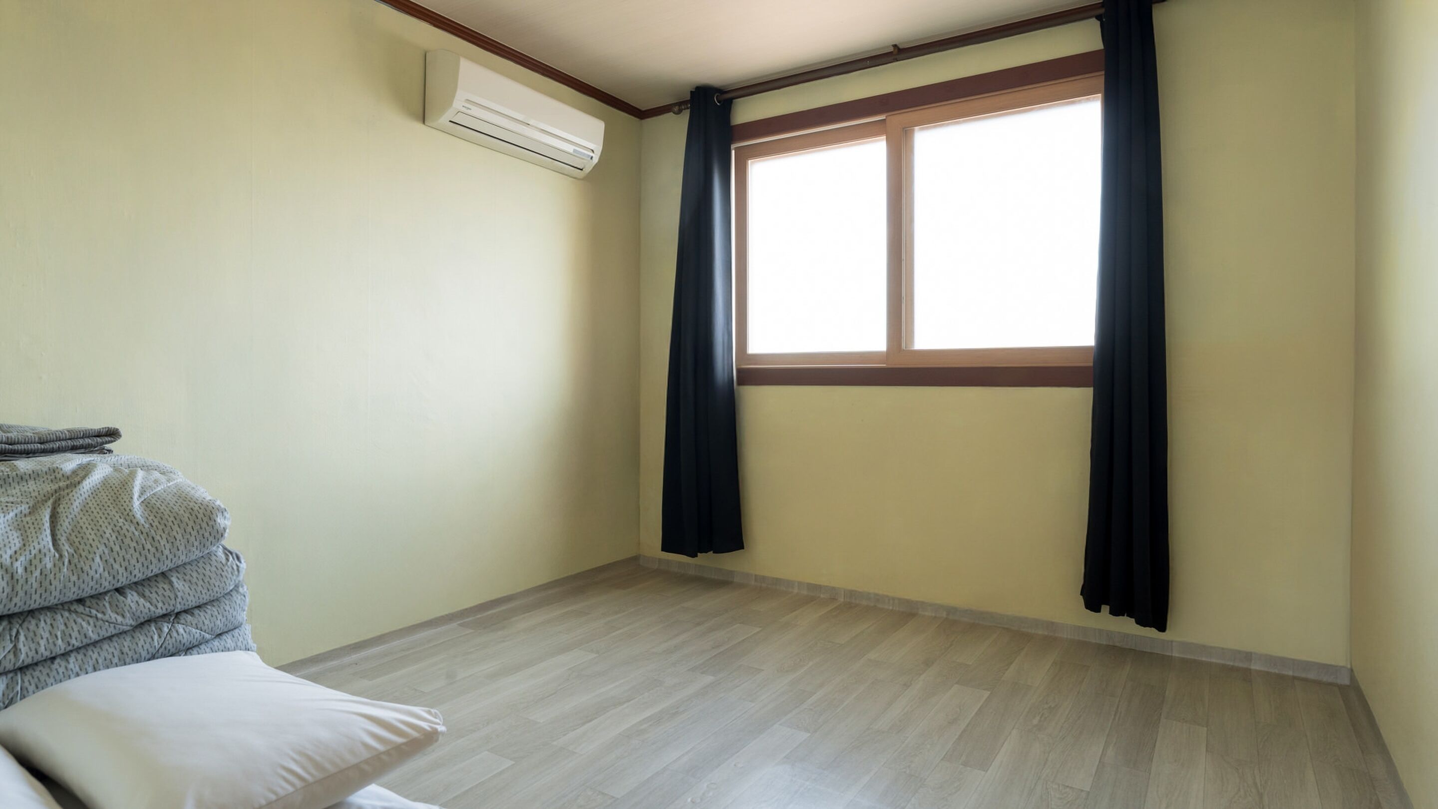A room with a large window, a ceiling-mounted air conditioner, and a bed with a pillow and a folded blanket.