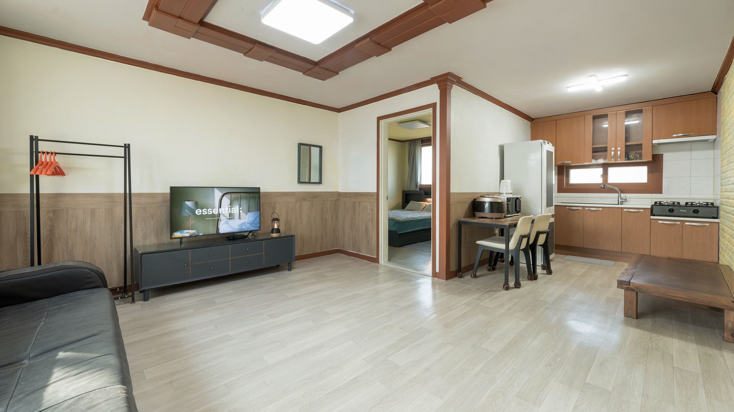 A modern living room with a flat-screen TV, a sofa, a dining area with chairs, and a kitchen with wooden cabinets and appliances.