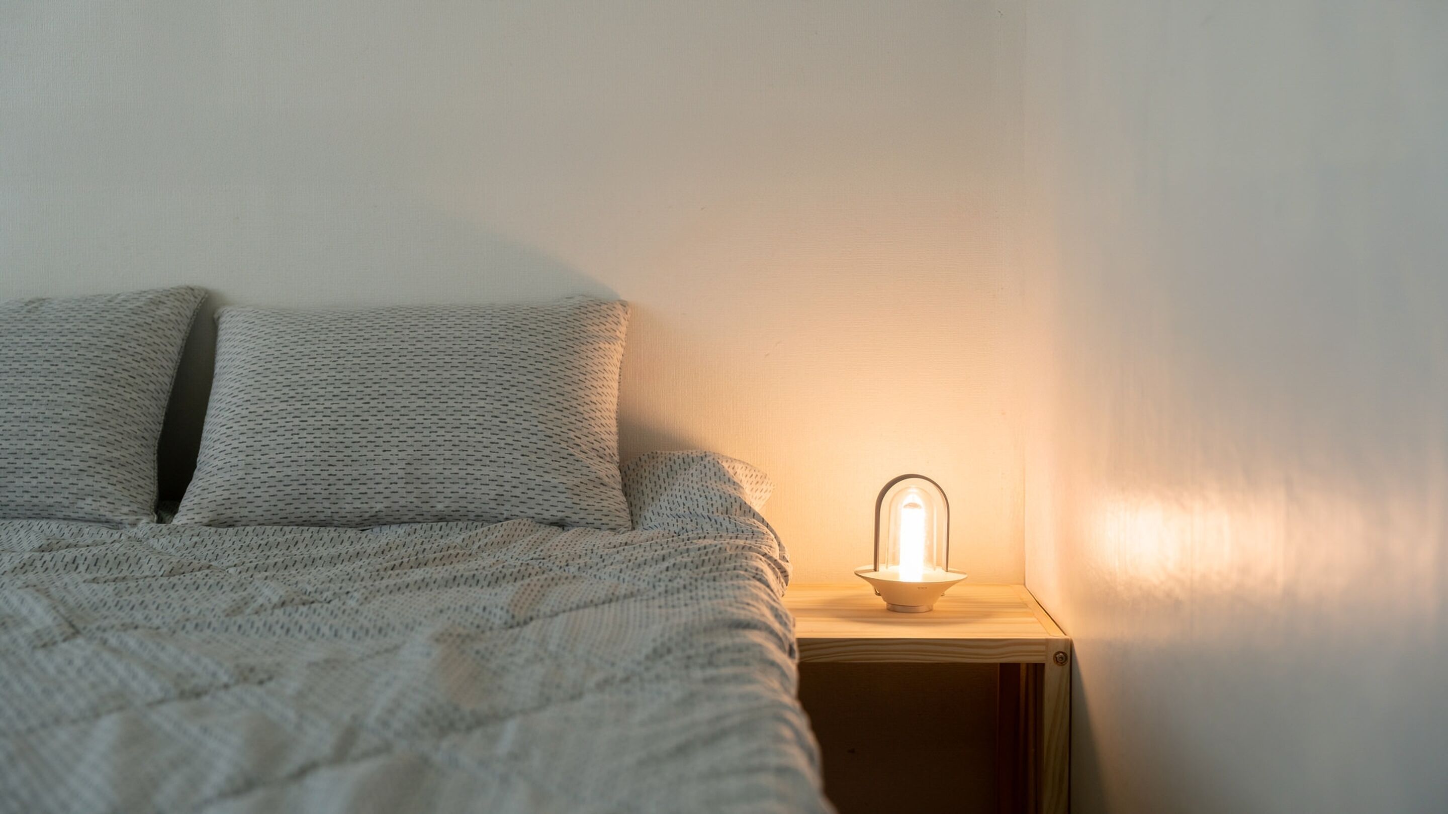 Una cama con un edredón tejido y almohadas, una mesita de noche con una lámpara y una pared lisa.