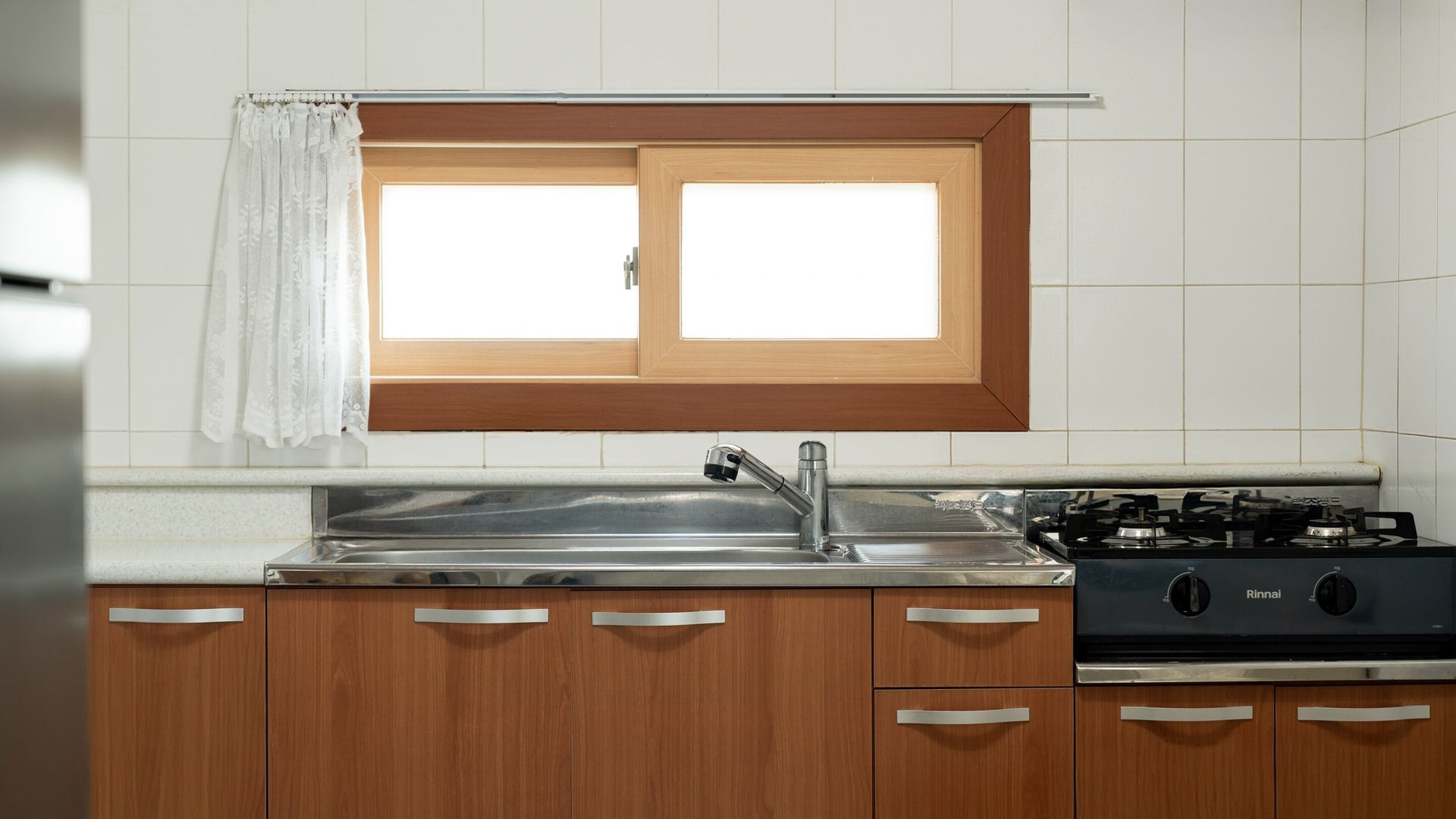 Una cocina con armarios de madera, un fregadero de acero inoxidable, una ventana con cortina y una estufa a gas.