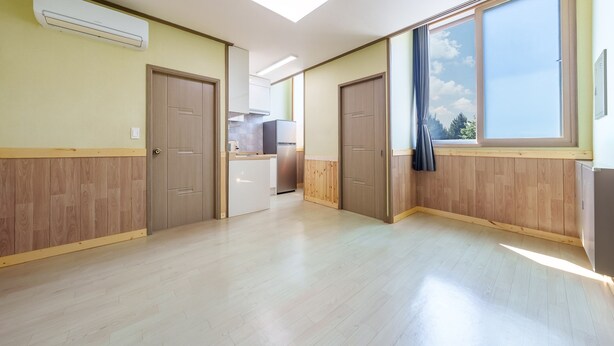 Un'ampia stanza con pavimento in legno, due porte, una finestra con vista e una piccola cucina sullo sfondo.