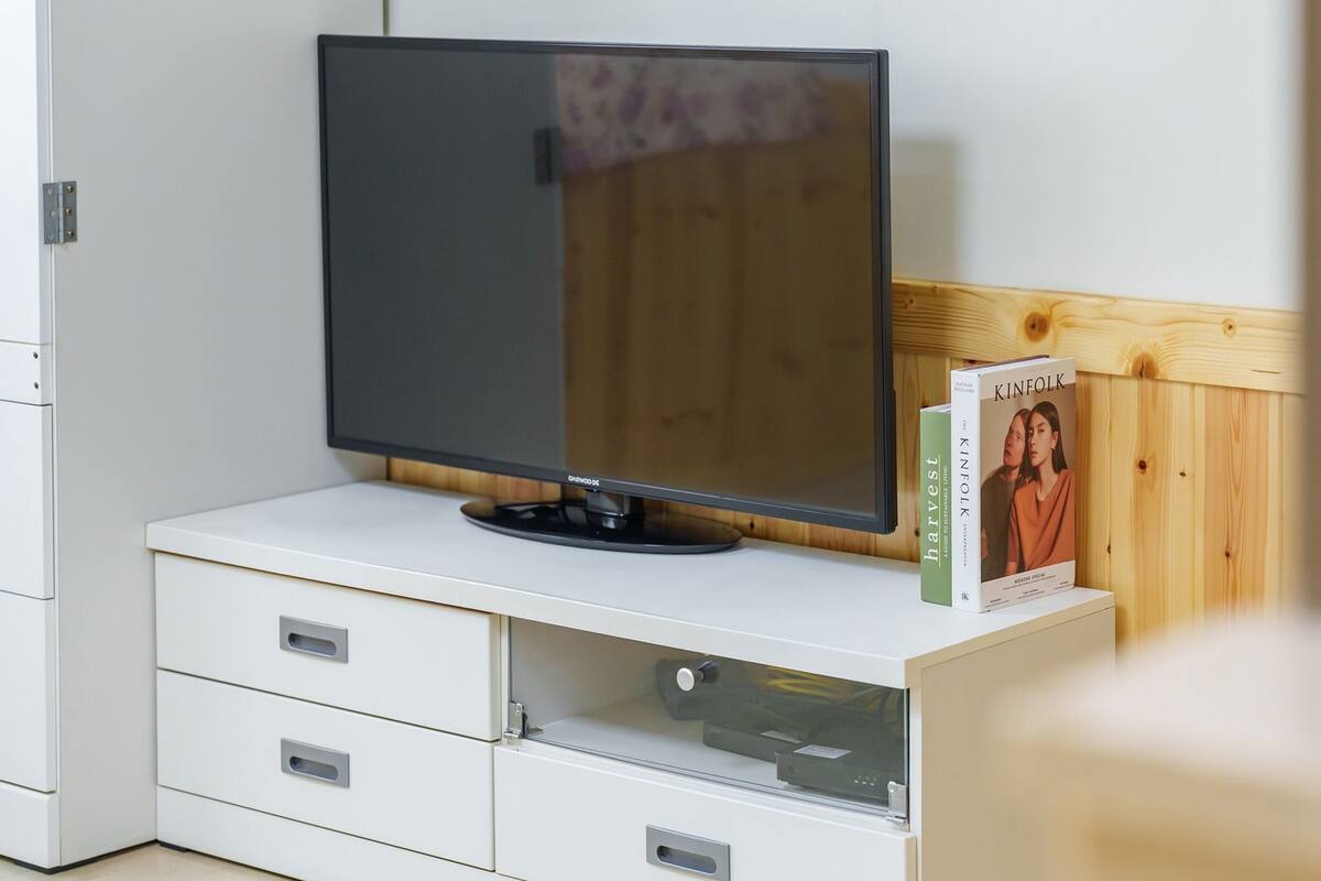A flat-screen TV on a white cabinet with drawers, a wooden panel wall, and a book titled 'KINFOLK' on the right.