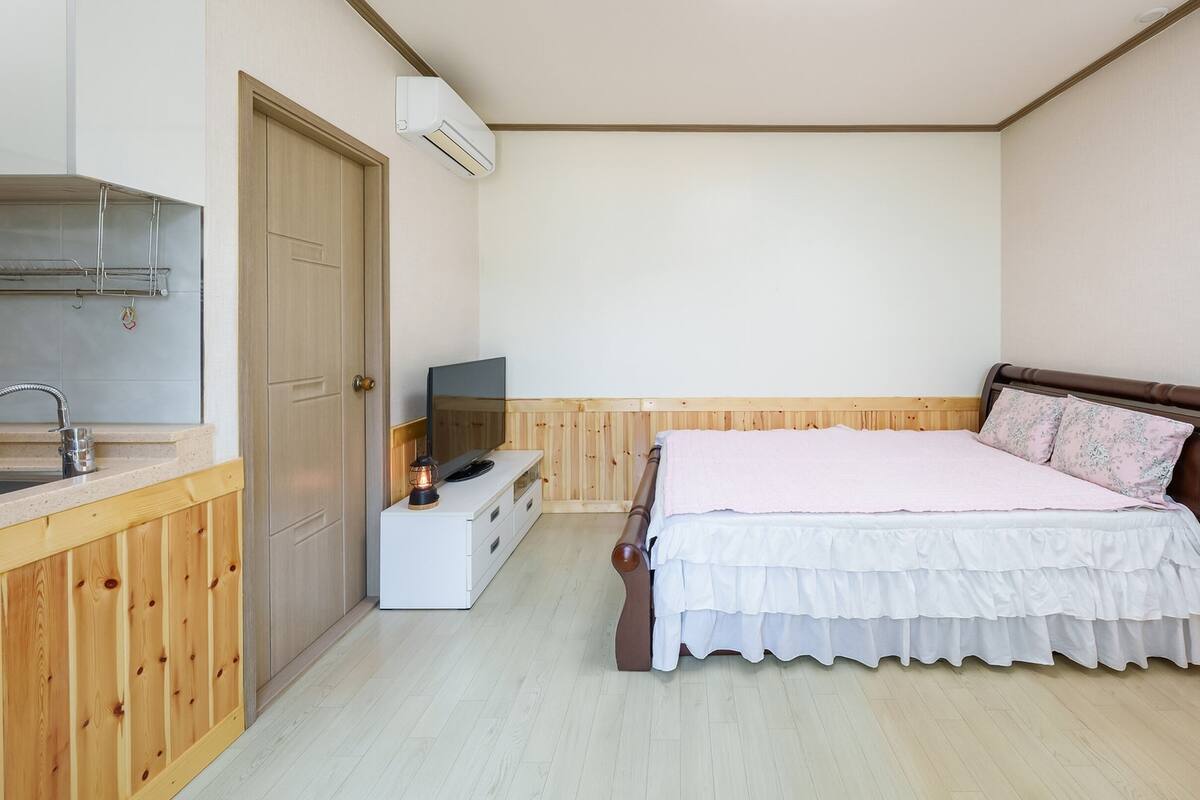 A bedroom with a wooden bed, a TV on a white dresser, a wooden wardrobe, and a kitchenette with a sink and an air conditioner.
