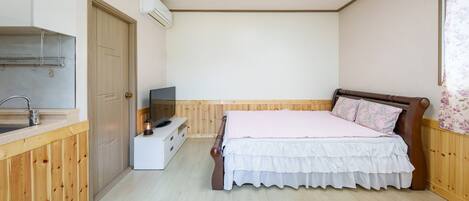 Un dormitorio con cama de madera, un televisor sobre un aparador blanco, un armario de madera y una kitchenette con lavamanos y aire acondicionado.