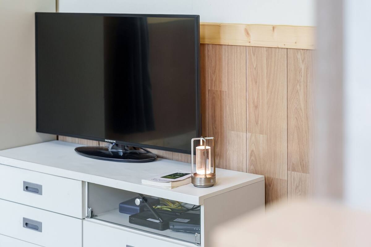 A modern room with a flat-screen TV on a white cabinet, a small table with a book and a candle, and a wooden panel wall.