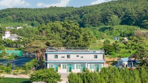 Un edificio a due piani con tetto piatto, circondato da vegetazione e una collina boscosa.