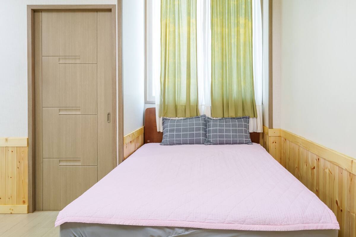 A single bed with a pink bedspread and two pillows, a wooden wardrobe, and a window with green curtains.