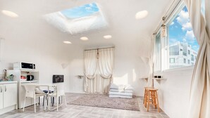 A modern, well-lit room with a skylight, a kitchenette, a dining area, and a large window offering a view of buildings. - Jeju Mulgogi Pension (Jeju City)