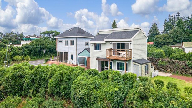 Une maison à deux étages dotée d’un balcon, entourée d’une végétation luxuriante et surplombant un ciel d’un bleu éclatant.