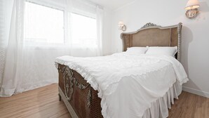 A neatly made bed with a white bedspread and pillows, a wicker headboard, and a window with sheer curtains.
