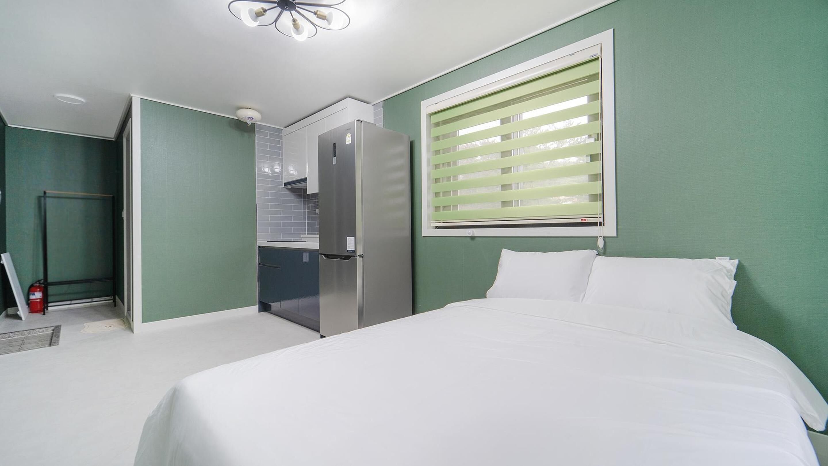 Une petite pièce comprenant un lit, une kitchenette et une fenêtre avec des stores.
