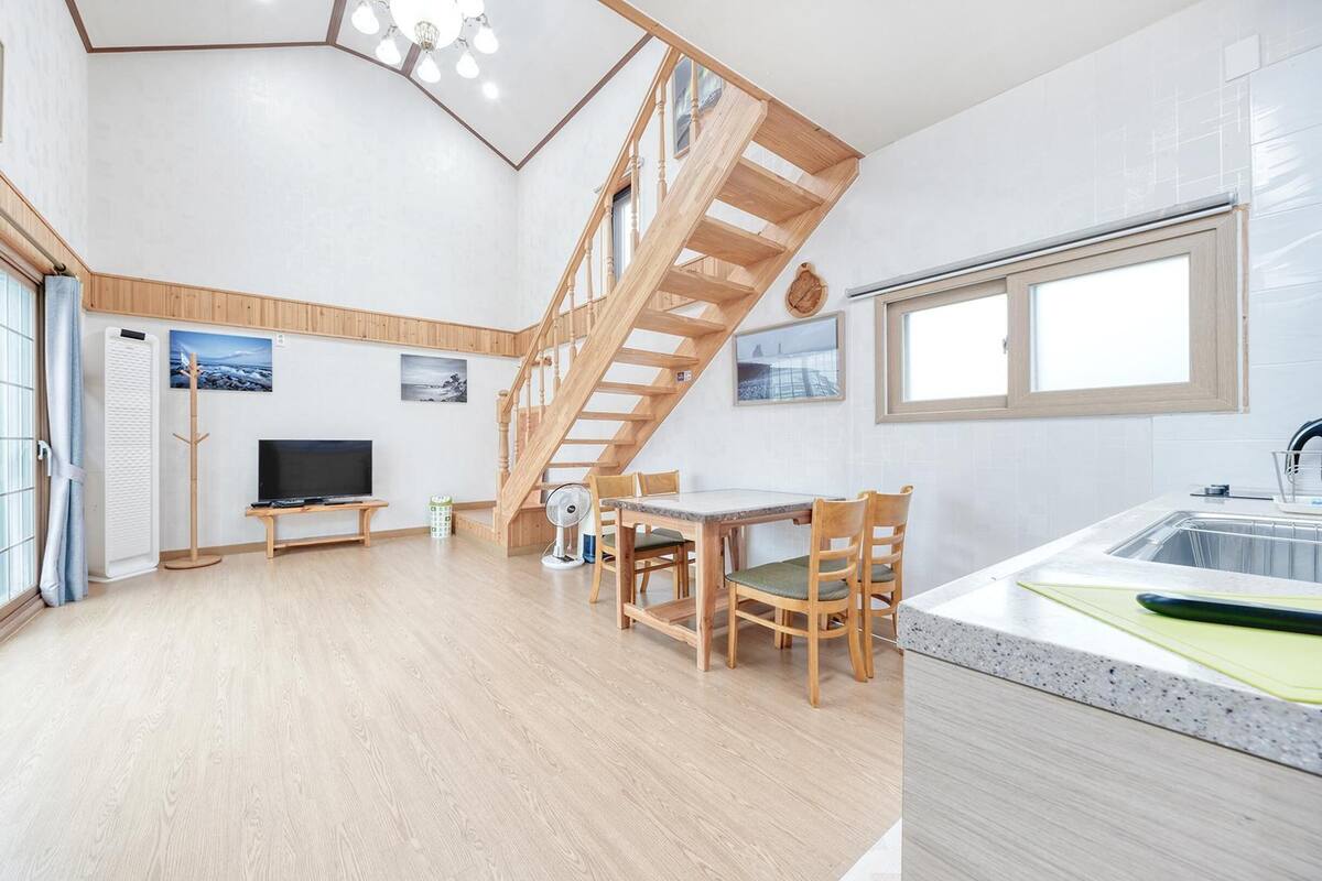 A spacious living area with a wooden staircase, a dining table with chairs, a flat-screen TV, and a kitchen with a sink and window.