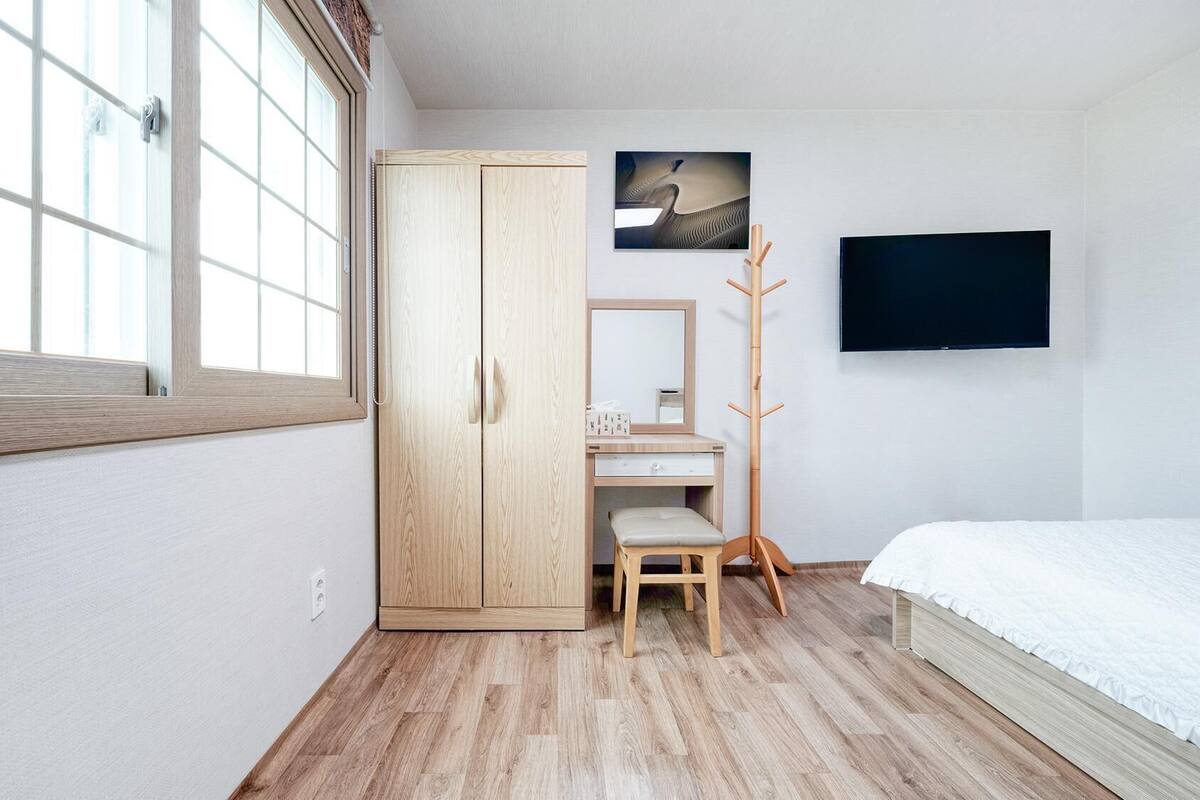 A bedroom with a wardrobe, a desk, a chair, a coat rack, a bed, and a TV.
