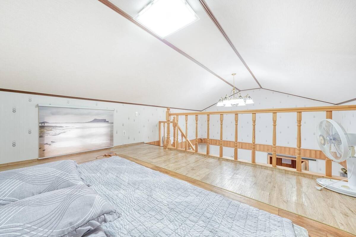 A room with a sloped ceiling, wooden flooring, a bed with a grey patterned bedspread, a standing fan, and a large window with a beach view.