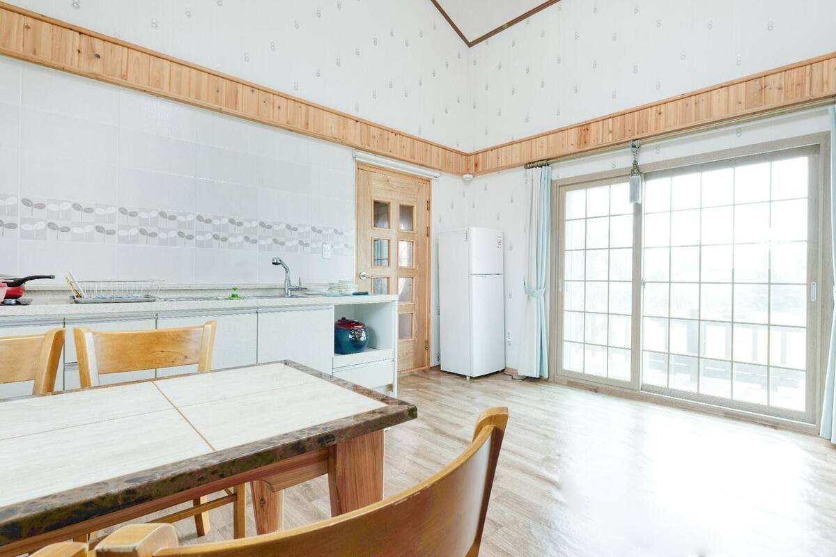 A modern kitchen with a dining area, wooden paneling, a refrigerator, and a large window with curtains.