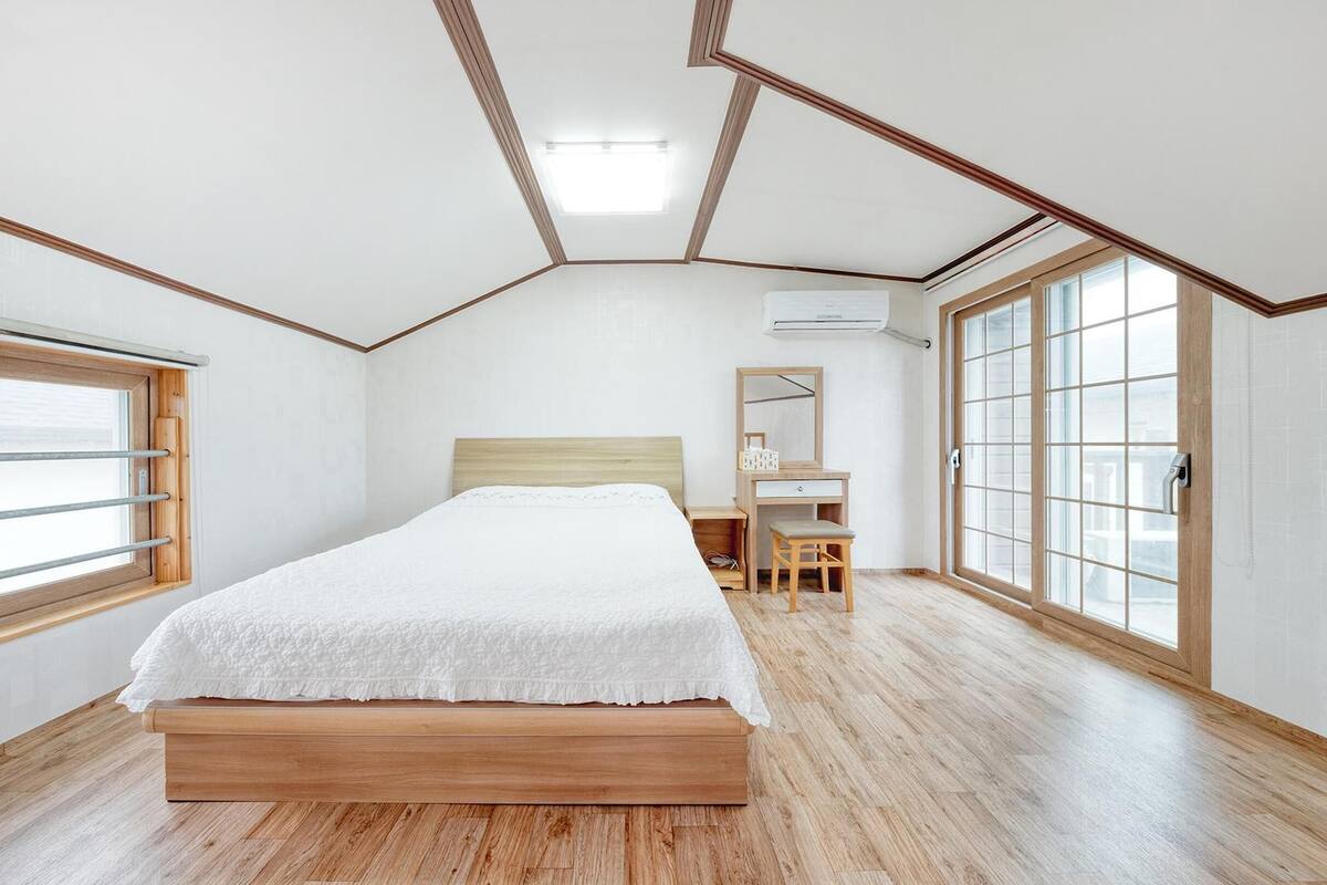 A bedroom with a wooden bed, a desk, and a chair, large windows, and an air conditioning unit.