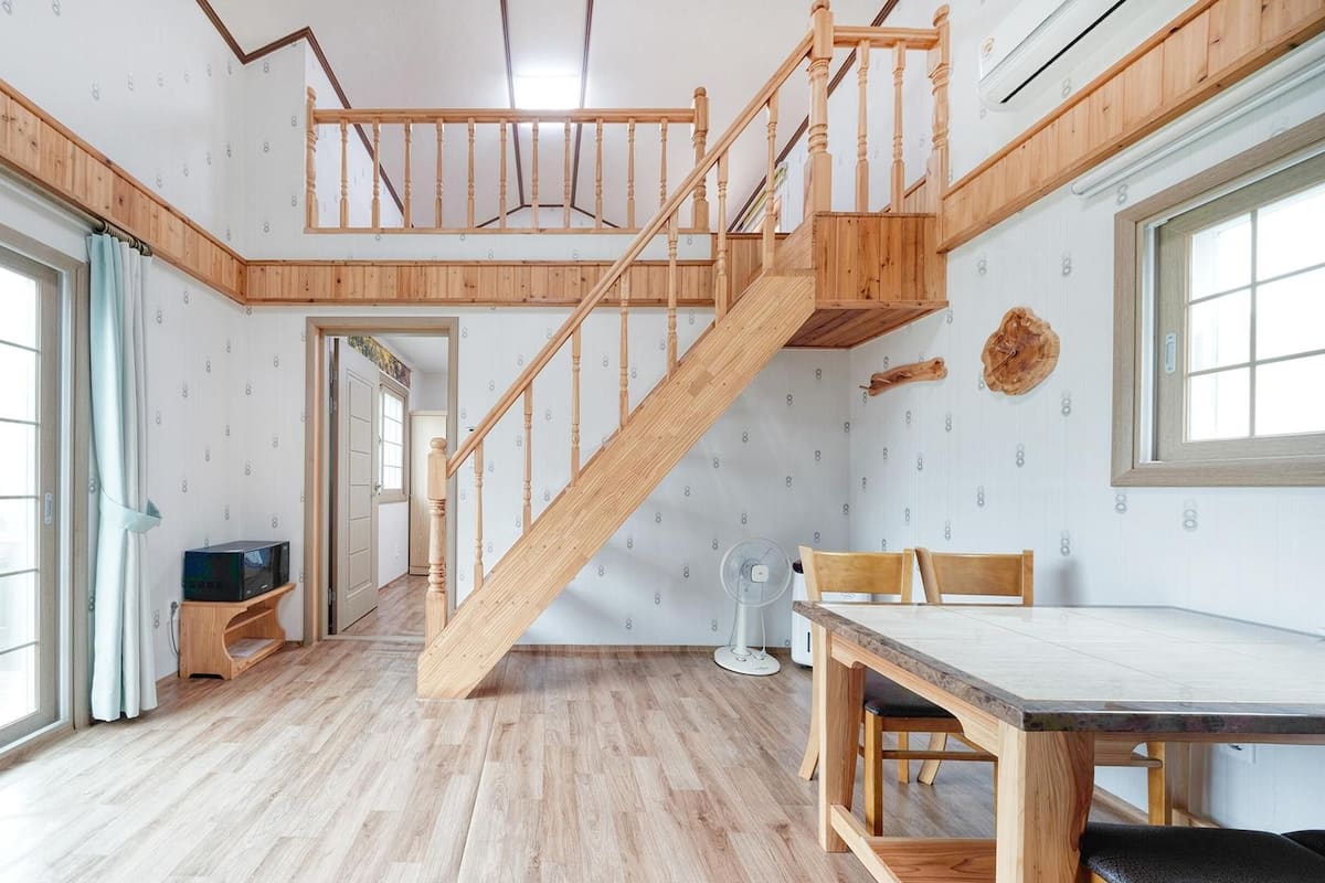 A spacious room with a wooden staircase, large windows, and a dining area.