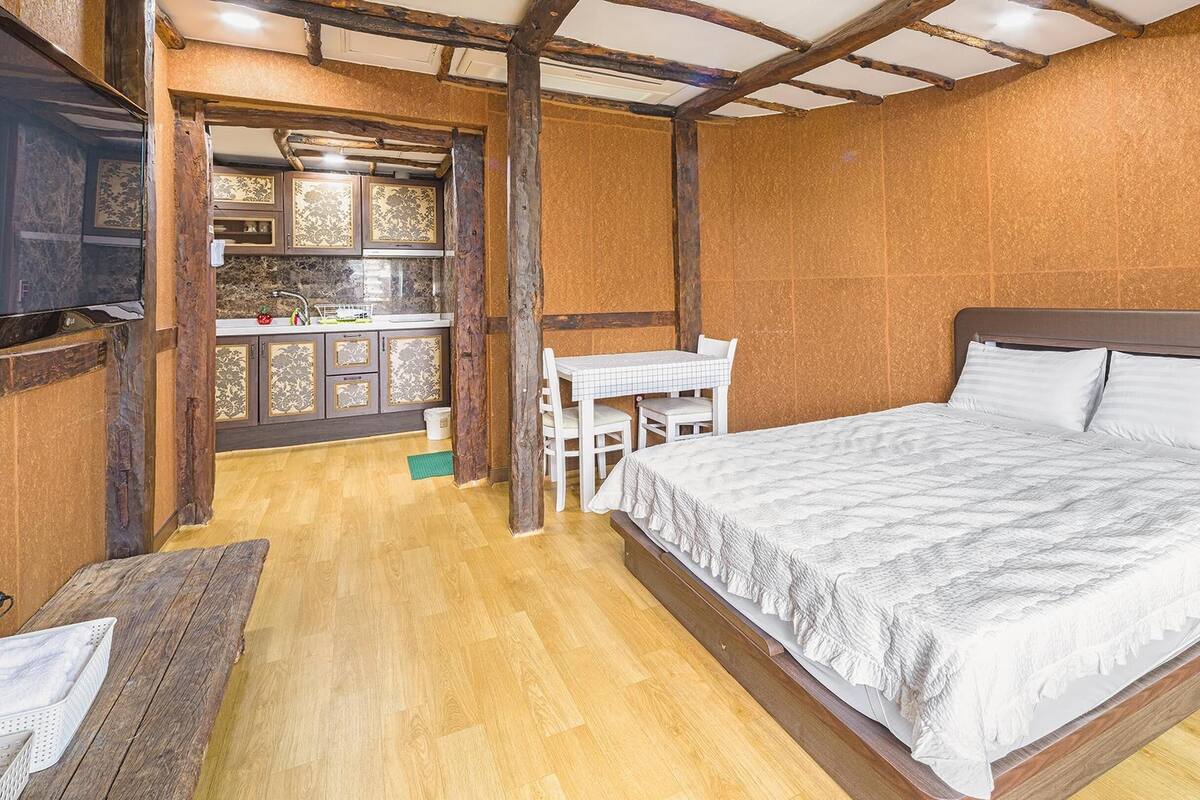 A bedroom with a wooden floor, a bed with white bedding, a small table with chairs, and a window with a view of the outside.