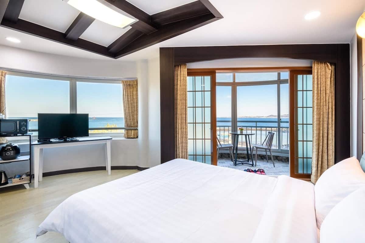 A hotel room with a bed, a desk with a TV, a view of the sea, and a balcony with chairs.