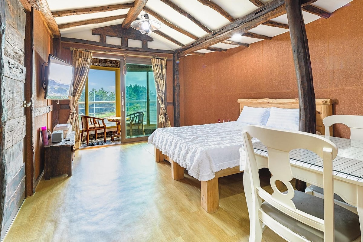 A rustic bedroom with a wooden bed, a small table, and a chair. There is a large window with curtains, and a view of greenery outside.