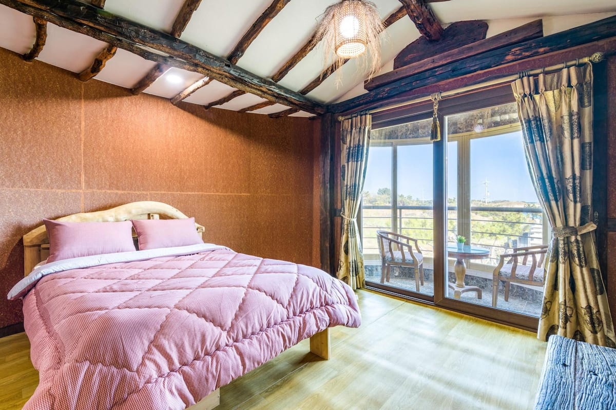 A bedroom with a large bed, wooden beams on the ceiling, and a view of the outdoors through a sliding glass door.