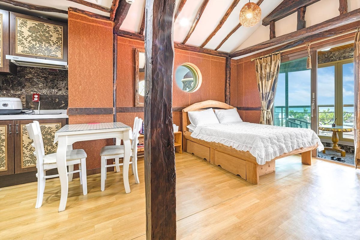 A bedroom with a wooden bed, a dining table with chairs, a kitchen area, and a balcony with a view.