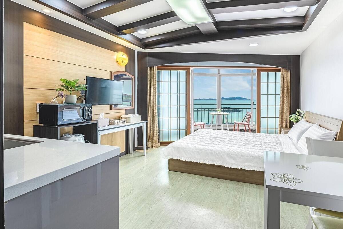 A modern hotel room with a bed, a desk with a TV, a balcony with a view of the sea, and a large window.