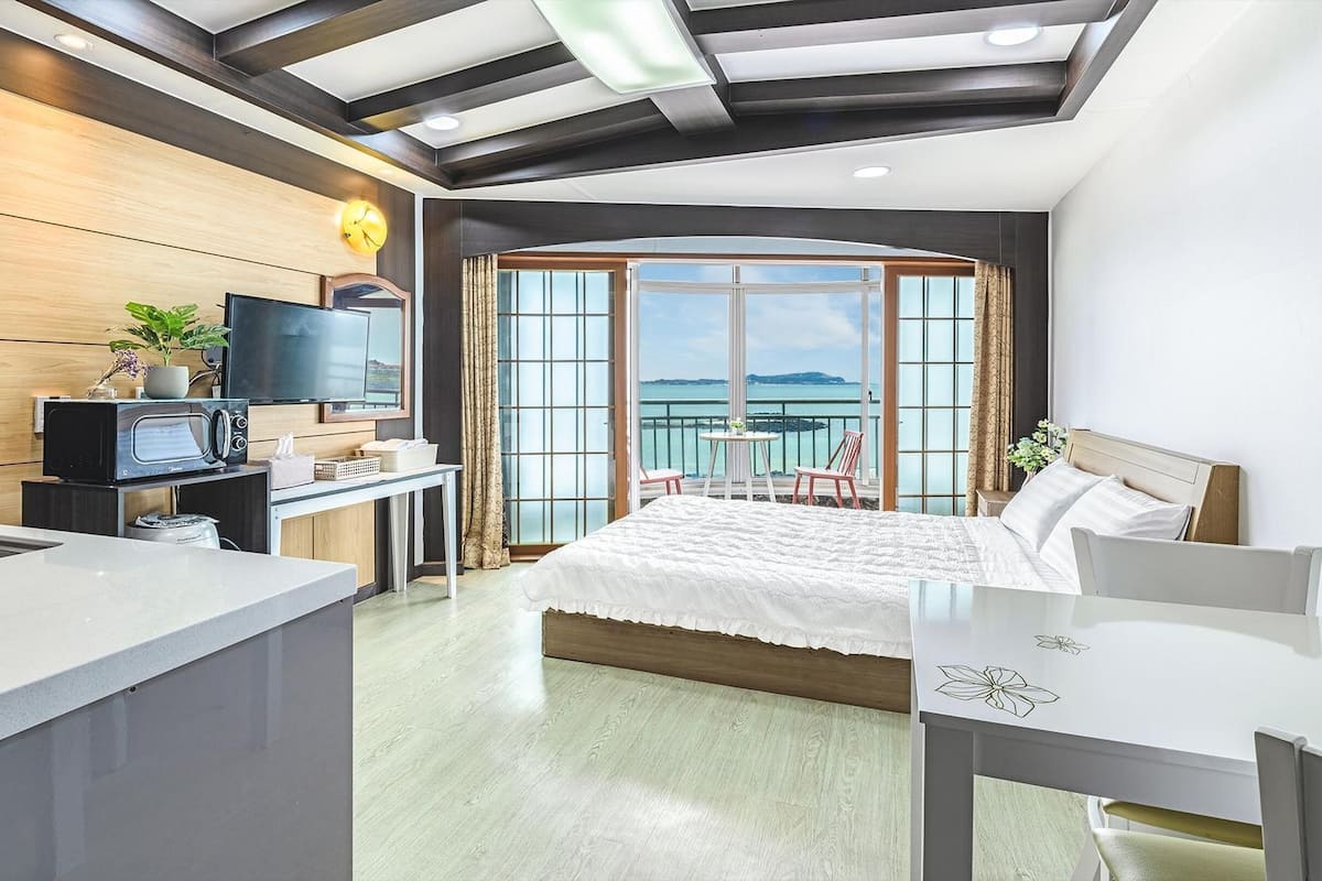 A modern hotel room with a balcony, a TV, a desk with a microwave, and a bed with a white duvet.