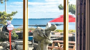A stone statue of a figure with a shovel, near a wooden bench and a red umbrella, overlooking a body of water with a rocky shoreline. - Jeju Padosoriminbak Pension (Jeju City)