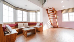 Um quarto amplo com área de estar com bancos de madeira, um mezanino com escada de madeira e grandes janelas que proporcionam iluminação natural.