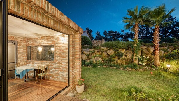 An open-air dining area with a brick wall, a wooden deck, and a stone wall with plants. - Seogwipo Hanonui Achim Pensiom (Seogwipo)