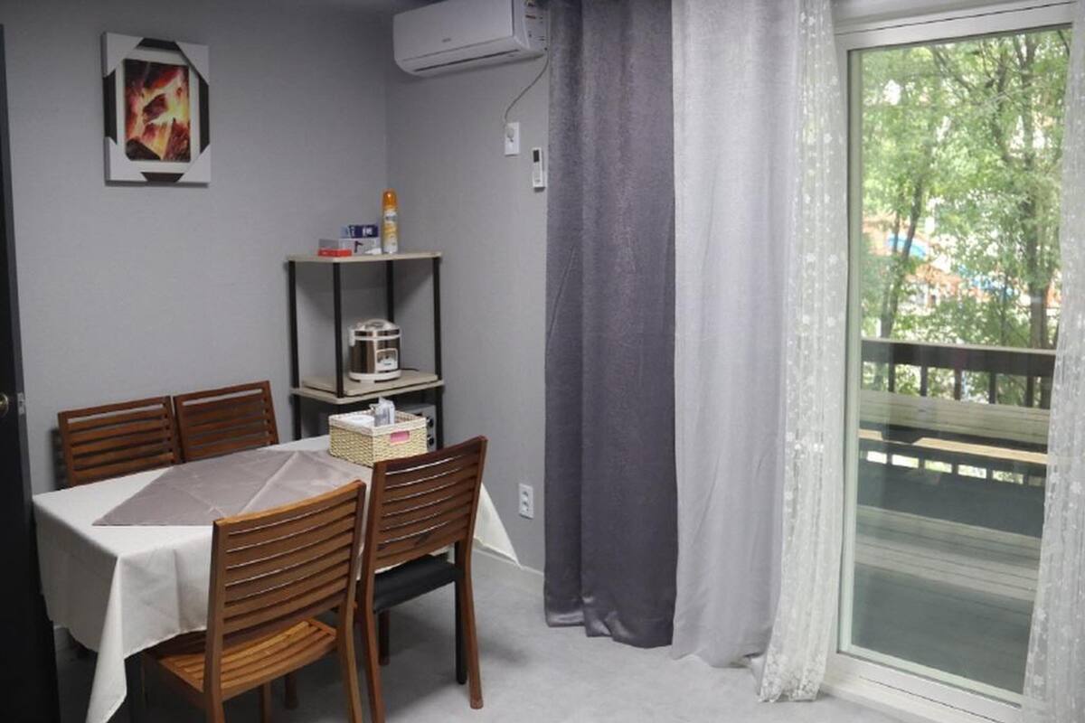 Ein Essbereich mit Tisch und Stühlen, einem Regal mit Gegenständen, einer Klimaanlage und Blick durch ein Fenster mit Vorhängen.