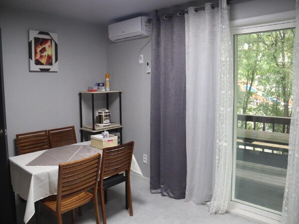 Ein Essbereich mit Tisch und Stühlen, einem Regal mit Gegenständen, einer Klimaanlage und Blick durch ein Fenster mit Vorhängen.