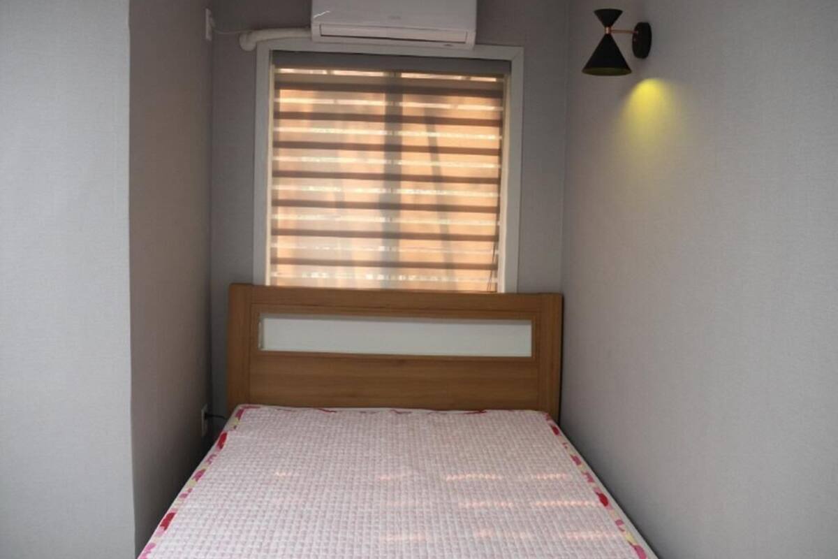 Ein kleines Zimmer mit einem Bett, einem Fenster mit Jalousien, einer Klimaanlage und einer Wandleuchte.